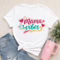 White t-shirt with 'Mama Vibes' design, paired with denim shorts and a pink tumbler on a neutral background.