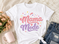 White t-shirt with 'Mama Mode' text on a fluffy surface with flowers and jeans.