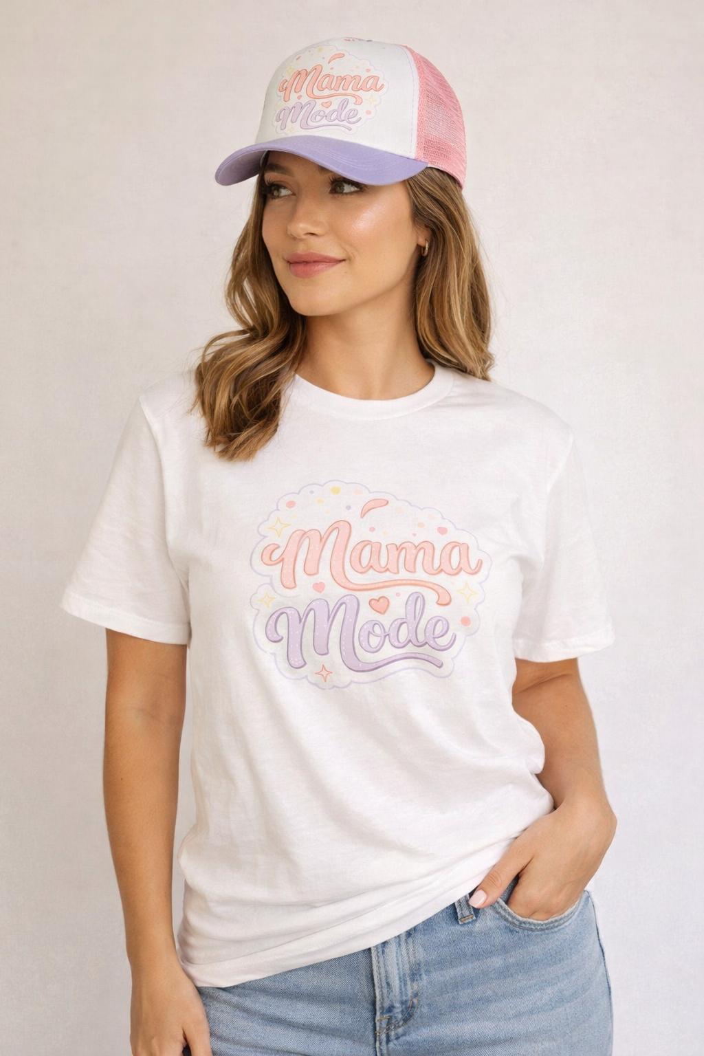 Woman wearing a white t-shirt with 'Mama Mode' text, light blue jeans, and a pink and white cap.